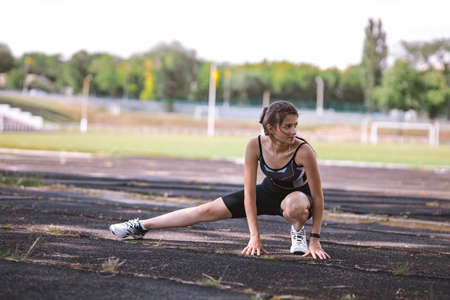 Caucasian beautiful girl athlete in dressed in sportswear with fitness tracker stretching and doing warm up exercise on city stadium in summer. Happy female outdoor sport and lifestyle healthy conceptの写真素材