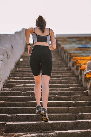 Beautiful Caucasian girl athlete dressed in sportswear: shorts and top, sneakers, fitness tracker runs on stadium stairs in summer. Happy young female outdoor sport, workout, healthy lifestyle conceptの写真素材