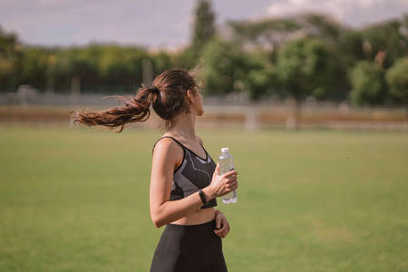 Beautiful Caucasian girl athlete in dressed in sportswear with fitness tracker holds plastic water bottle in her hand on stadium in hot summer. Happy female outdoor sport, lifestyle healthy conceptの写真素材