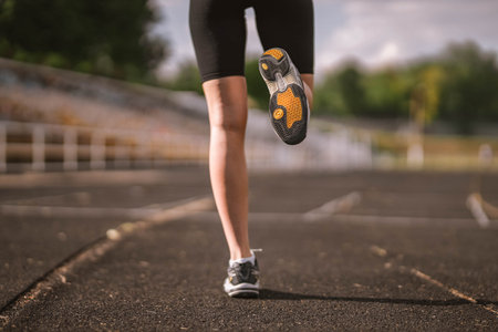 Close-up photo of the Caucasian athlete's leg dressed in sportswear: shorts and top, sneakers with fitness tracker starting running on track at stadium in summer. Happy female outdoor sport conceptの写真素材
