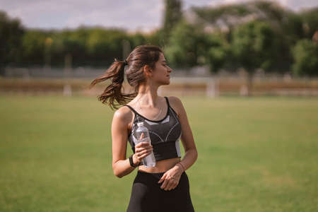 Beautiful Caucasian girl athlete in dressed in sportswear with fitness tracker holds plastic water bottle in her hand on stadium in hot summer. Happy female outdoor sport, lifestyle healthy conceptの写真素材