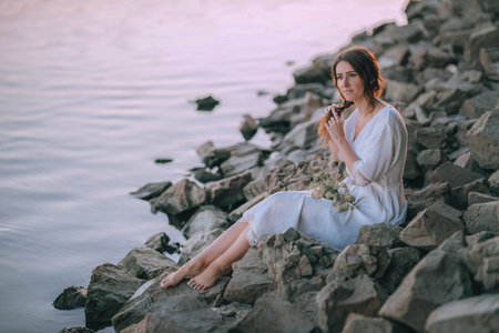 Sensual girl in white long vintage dress makes a braid near the river on stones. Harmony and rest with nature. Ukrainian tradition celebration of Ivana Kupala. Relax, travel, quiet placeの写真素材