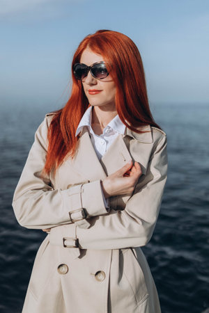 Stylish red hair lady stands, posing on the seaside in the afternoon, long brown trench-coat, sunglassesの写真素材