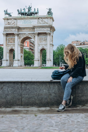 Milan, Italy- May 13, 2023: Girl sits near Arch of Peace or Sempione Gate in Milan, near Sempione Park. Exterior view of the marble gate of the Arco della Sempione, a peace memorial in Milan.の写真素材