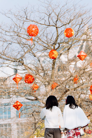Two Women Walking Under Tree with Red Lanterns Hanging in Winter Landscapeの写真素材