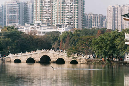 Stone Arch Bridge Over Calm Lake with Urban Buildings and Trees in Backgroundの写真素材