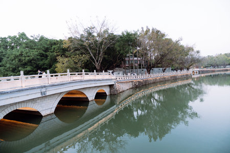 Stone arch bridge spans calm river with tree-lined banks and reflective water under overcast skyの写真素材