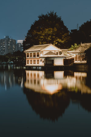 Traditional Chinese Pavilion Illuminated at Night Reflecting on Calm Lake with City Skyline in Backgroundの写真素材