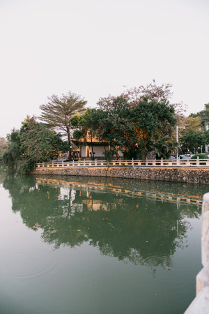 Traditional building beside calm river with tree reflections and stone railing under overcast skyの写真素材