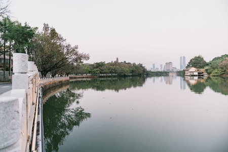 City River Scene with Stone Bridge, Trees, and Distant Skyscrapers Reflecting in Calm Waterの写真素材
