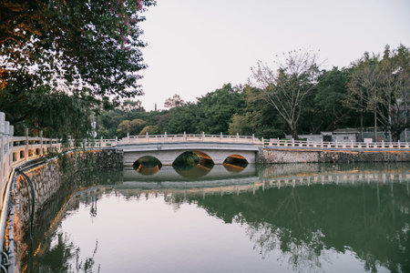 Ancient stone bridge with three arches spans calm river, surrounded by trees and reflective water under soft daylightの写真素材