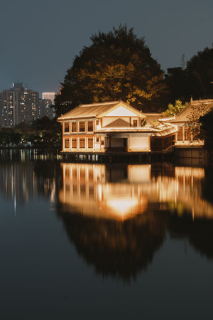 Traditional Chinese Pavilion Illuminated at Night with Perfect Reflection on Calm Lake Near City Skylineの写真素材