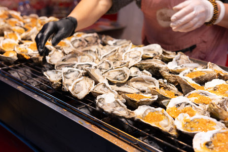 Person grilling oysters on barbecue grill at food market with gloved hands and toppingsの写真素材