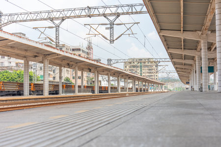 Empty train station platform with overhead electric lines, concrete pillars, and urban buildings in background under overcast skyの写真素材