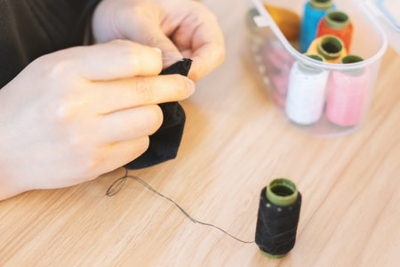 Hands sewing black fabric with needle and thread on wooden table near spools of colorful thread