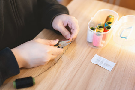 Person sewing with needle and thread at wooden table, crafting supplies nearby