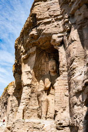 Ancient Buddha statue carved into cliff face under blue sky, weathered stone sculpture in historic cave templeの写真素材
