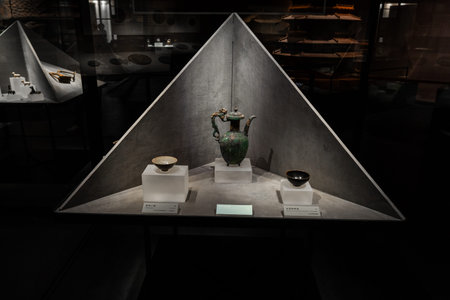 Ancient ceramic teapot and bowls displayed under triangular glass case in museum exhibitの写真素材