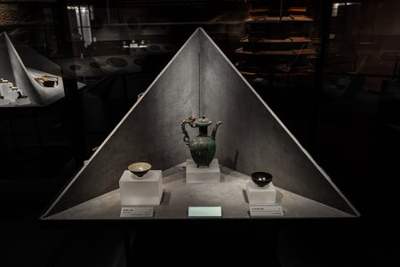Ancient ceramic vase and bowls displayed in triangular museum exhibit under focused lightingの写真素材