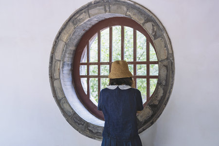 Woman in straw hat looking through circular window with wooden lattice, white wall backgroundの写真素材