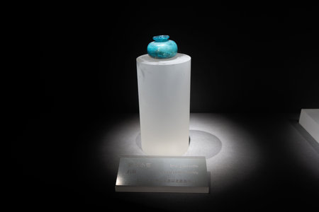 Ancient turquoise glass vessel displayed on white pedestal under spotlight in museum setting