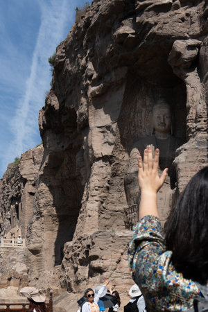 Tourist reaching hand toward ancient stone Buddha statue carved into cliff face under blue skyの写真素材