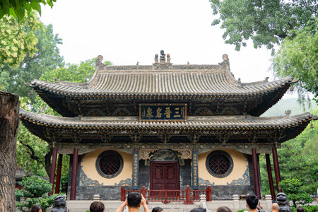 Traditional Chinese Temple with Gray Tiled Roof and Red Doors, Surrounded by Green Trees, Visitors...