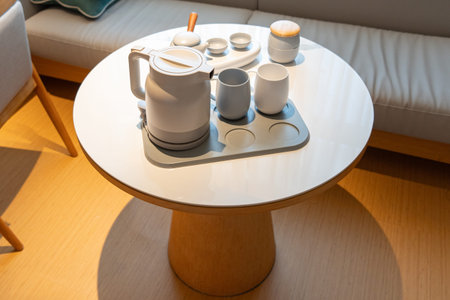 Modern white electric kettle and ceramic tea set on round wooden table in cozy living roomの写真素材