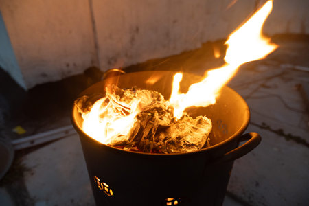 Burning wood fire in black metal pot with flames rising, outdoor setting at night, rustic heating...