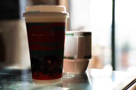 Disposable coffee cup with red design and ceramic mug on glass table near windowの写真素材