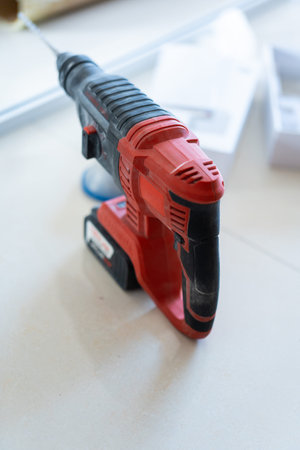 Red and black cordless hammer drill on white surface with construction tools in background, studio
