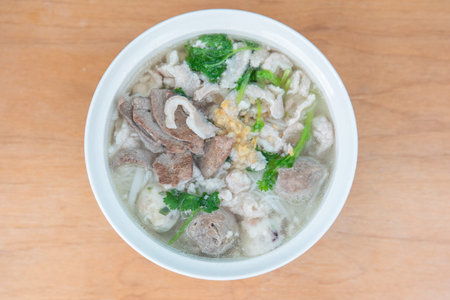Steaming bowl of Chinese pork noodle soup with greens and meatballs, served on wooden table in