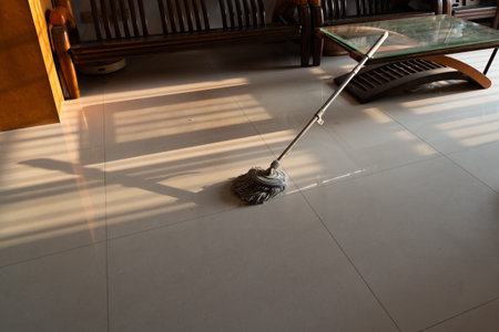 Mop resting on tiled floor in sunlit living room with wooden furniture and glass table, typical