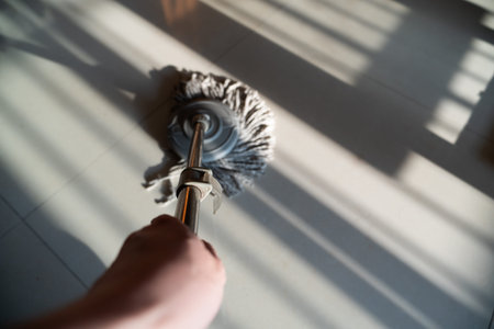 Hand holding mop cleaning tiled floor with sunlight shadows in modern home interior