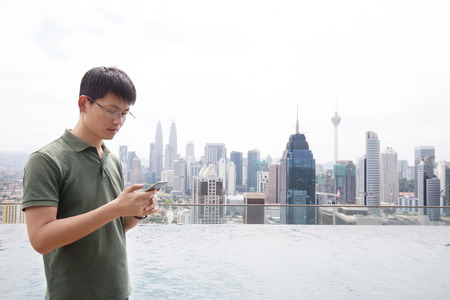 young chinese man with mobile phone stands by swimming pool on balcony in midtown of kuala lumpur in cloud skyのeditorial素材