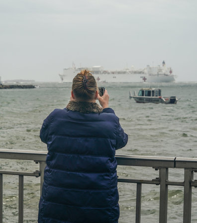 NEW YORK - APRIL 30, 2020: Spectators on the Brooklyn shoreline take pictures as the USNS Comfort...