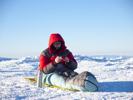 A man sits in a sleeping bag and drinking tea from a bottle on the background of the winter mountains.の写真素材