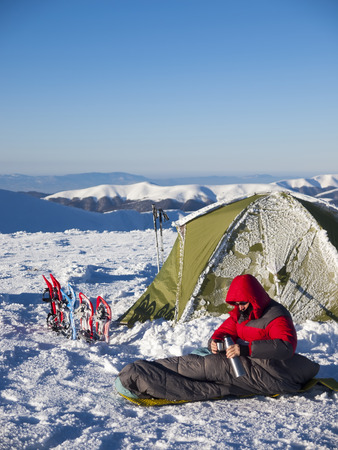 A man sits in a sleeping bag near the tent and snowshoes and drinking tea from a thermos on the background of the winter mountains.の写真素材