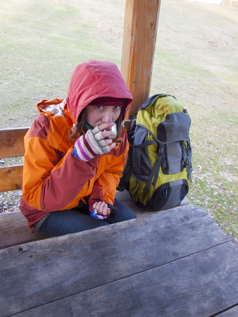 Girl with a backpack sitting at a wooden table and drinking from a thermos.の写真素材