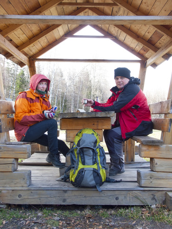 Man and woman sitting in a wooden gazebo in the woods and drink from a thermos hot drink.の写真素材