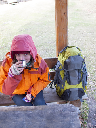 Girl with a backpack sitting at a wooden table and drinking from a thermos.の写真素材