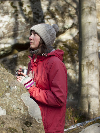 A young girl stands in the forest among the rocks with a warm drink in a mug.の写真素材