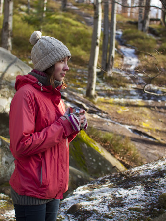 A young girl stands in the forest among the rocks with a warm drink in a mug.の写真素材