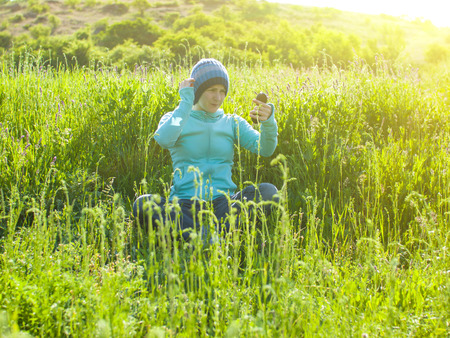 A young girl in a knitted hat sitting on green grass in a field and looks in the mirror.の写真素材