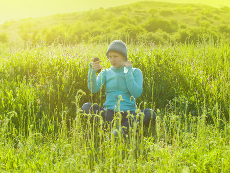 A young girl in a knitted hat sitting on green grass in a field and looks in the mirror.の写真素材
