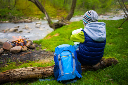 The child who is traveling is looking at a map sitting near a fire in a clearing.の写真素材