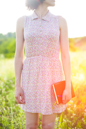 Girl in summer dress holding a book standing on the Sunny meadow. Soft focus.の写真素材