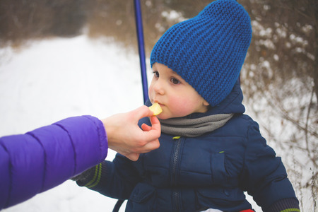 the little boy in the blue knit cap sitting on a sled, and eating an apple from mother's hands.の写真素材