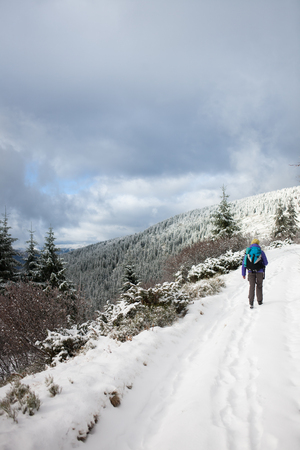 a woman walks and leaves footprints in the snow, young woman with a backpack walking along the trail in the mountains on the background of snowy mountains and cloudy sky.の写真素材