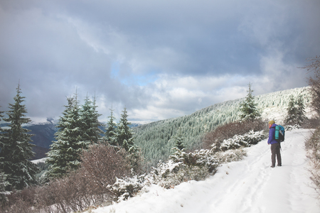 a woman walks and leaves footprints in the snow, young woman with a backpack walking along the trail in the mountains on the background of snowy mountains and cloudy sky.の写真素材